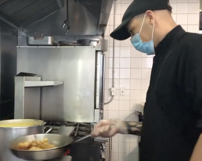Cook with saute pan in New York City catering kitchen wearing ppe mask during COVID-19 crisis.